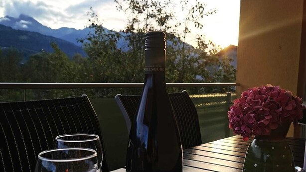 Una botella de vino sobre una mesa con dos copas, un jarrón con flores y una vista panorámica de montañas al fondo.