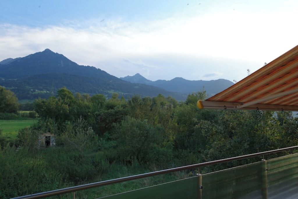 Blick von einem Balkon mit Metallgeländer auf eine gebirgige Landschaft mit grüner Vegetation und einem kleinen Bauwerk in der Ferne.