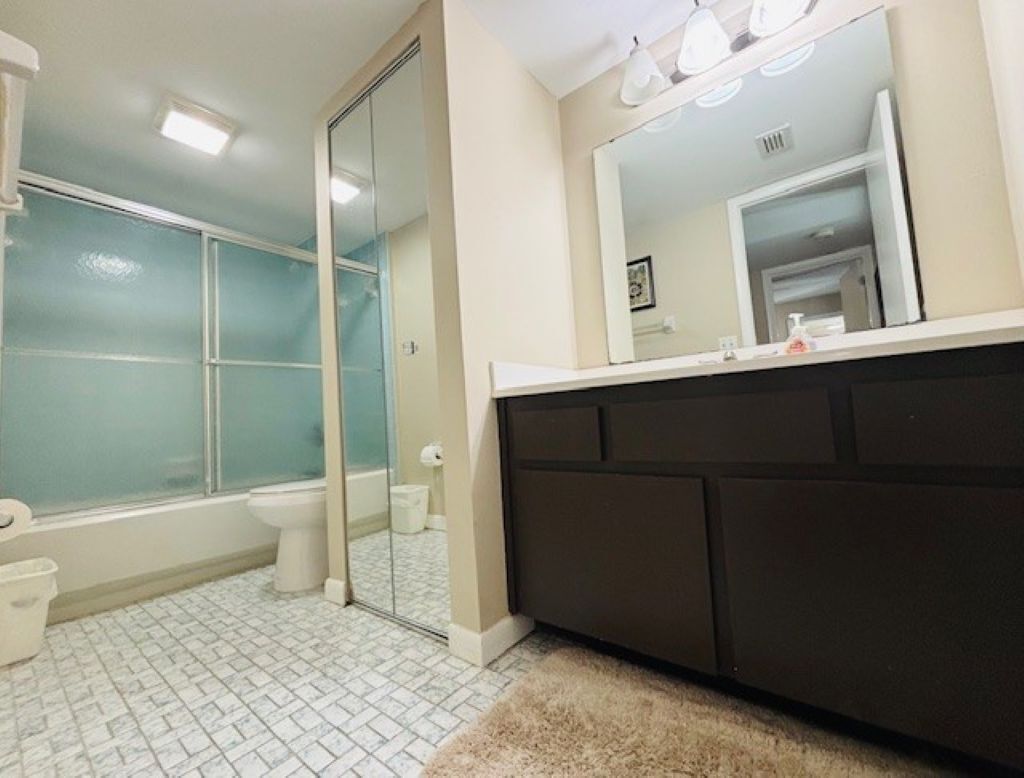 Une salle de bain avec une douche à parois de verre, des toilettes, un lavabo avec un miroir et un tapis blanc sur le sol.