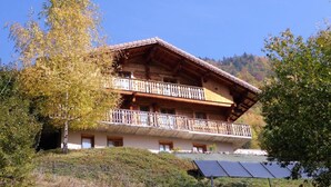 A two-story wooden chalet with balconies, set against a mountainous backdrop.