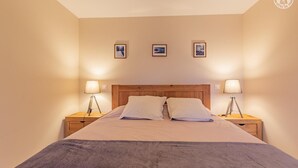 A neatly made bed with two pillows, flanked by two bedside tables with lamps, in a room with plain walls and three framed pictures above the bed.