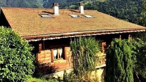A wooden cabin with a sloped roof, two chimneys, and a window with curtains.