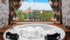 A bathroom with a large bathtub filled with bubbles, a wooden vanity, and a view of hot air balloons and a mountain through a window with curtains. - Cappadocia Fiesta Stone House (Nevsehir)