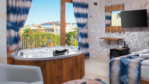 A room with a stone wall, a wooden bathtub, a bed with a blue and white patterned bedspread, a flat-screen TV mounted on the wall, and a balcony with a view of houses. - Cappadocia Fiesta Stone House (Nevsehir)
