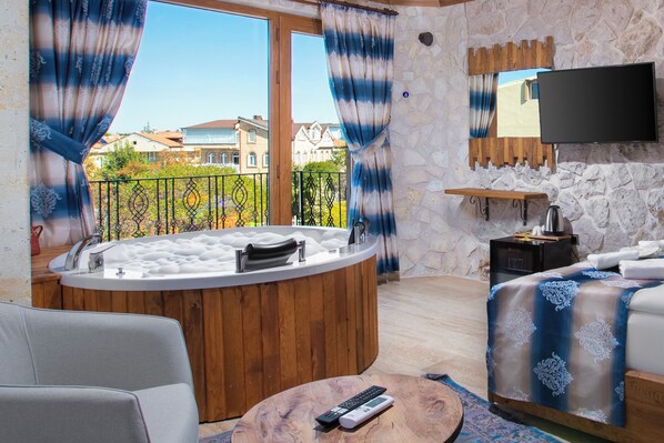 A room with a stone wall, a wooden bathtub, a bed with a blue and white patterned bedspread, a flat-screen TV mounted on the wall, and a balcony with a view of houses.