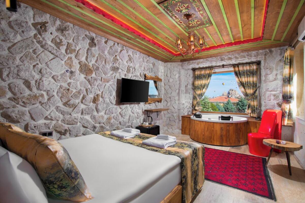 A room with a stone wall, a bed with a red and black patterned rug, a wooden ceiling with a chandelier, a red chair, and a view of a mountain through a window.