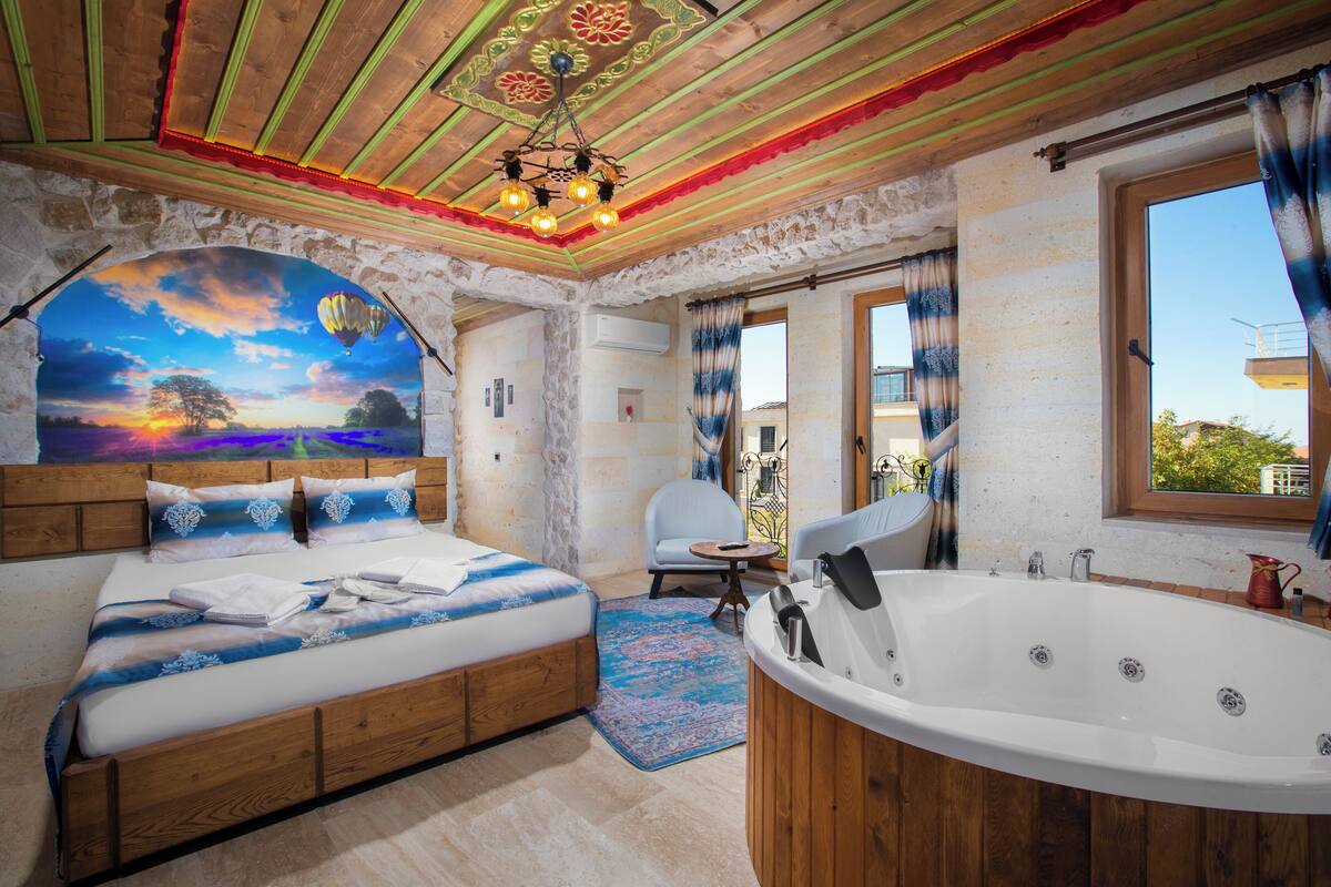 A bedroom with a large bed, a wooden headboard, a ceiling with a painted design, a wall mural, a bathroom with a bathtub, and a window with curtains.
