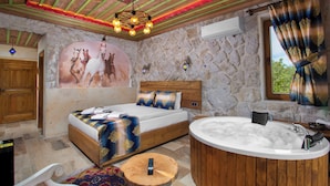 A bedroom with a large bed, a wooden headboard, a stone wall, a large window with curtains, a wooden side table, and a bathtub. - Cappadocia Fiesta Stone House (Nevsehir)