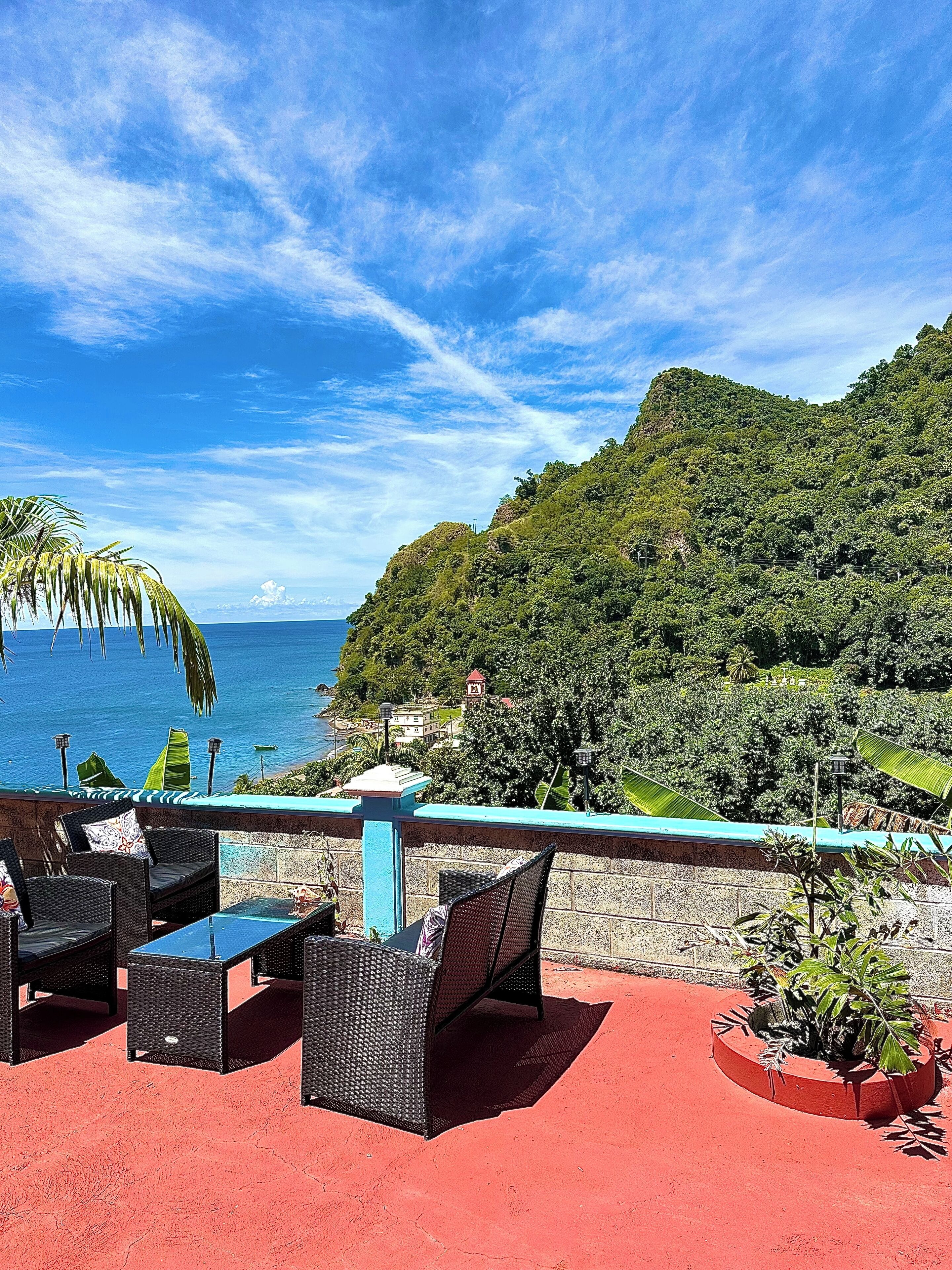 A terrace with wicker furniture, a red floor, and a view of the ocean and a hillside.