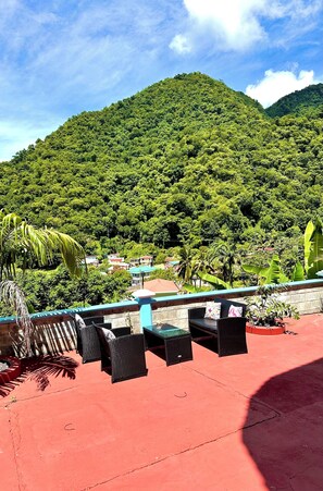 A terrace with wicker furniture and a red floor, overlooking a lush green mountain and a village below.