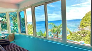A room with a large window overlooking a coastal view, a striped sofa, and a small table. - The Big Blue Condo (Soufriere)