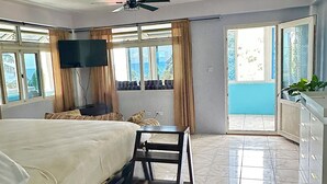 A hotel room with a bed, a TV mounted on the wall, a ceiling fan, and a view of the outdoors through large windows. - The Big Blue Condo (Soufriere)