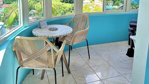 A corner room with a round table and two wicker chairs, large windows with a view of a green landscape, and a tiled floor. - The Big Blue Condo (Soufriere)
