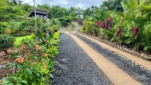 Un sentier bordé de gravier et agrémenté de plantes tropicales, qui mène à un bâtiment doté d’un toit en pente.