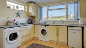 A compact kitchen with a washing machine, dishwasher, and a window with curtains. - Marrams View | Winterton Cottages (Winterton On Sea)