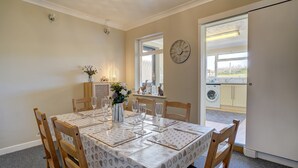 A dining area with a table set for four, wooden chairs, a vase with flowers, and a clock on the wall. - Marrams View | Winterton Cottages (Winterton On Sea)