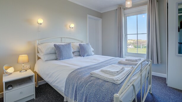 A neatly made bed with a white and blue striped blanket, a bedside lamp, and a small drawer unit. - Marrams View | Winterton Cottages (Winterton On Sea)