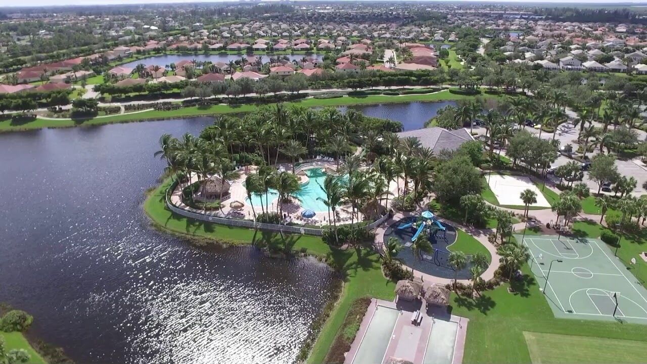 An aerial view of a residential area with a large swimming pool surrounded by palm trees, a basketball court, and a tennis court.