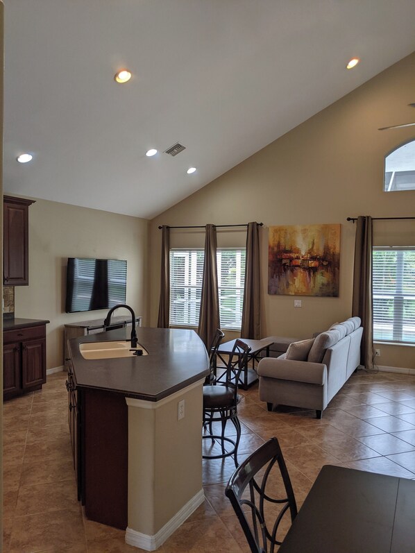 A spacious kitchen with a central island, dining area with a sofa and chairs, and a large window with blinds.