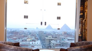 Ett rum med ett stort fönster som ger utsikt över en stadssiluett och pyramider. I förgrunden står två stolar och ett litet bord.