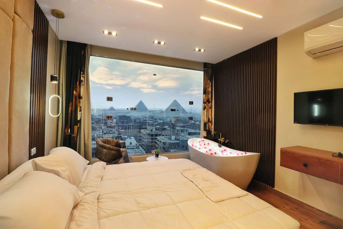 A hotel room with a large bed, a bathtub with rose petals, a television, and a view of a cityscape with a pyramid.