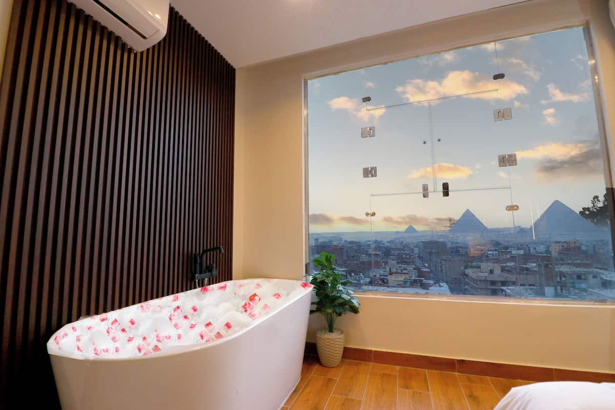 A modern bathroom with a large bathtub, a potted plant, and a cityscape view through a large window.