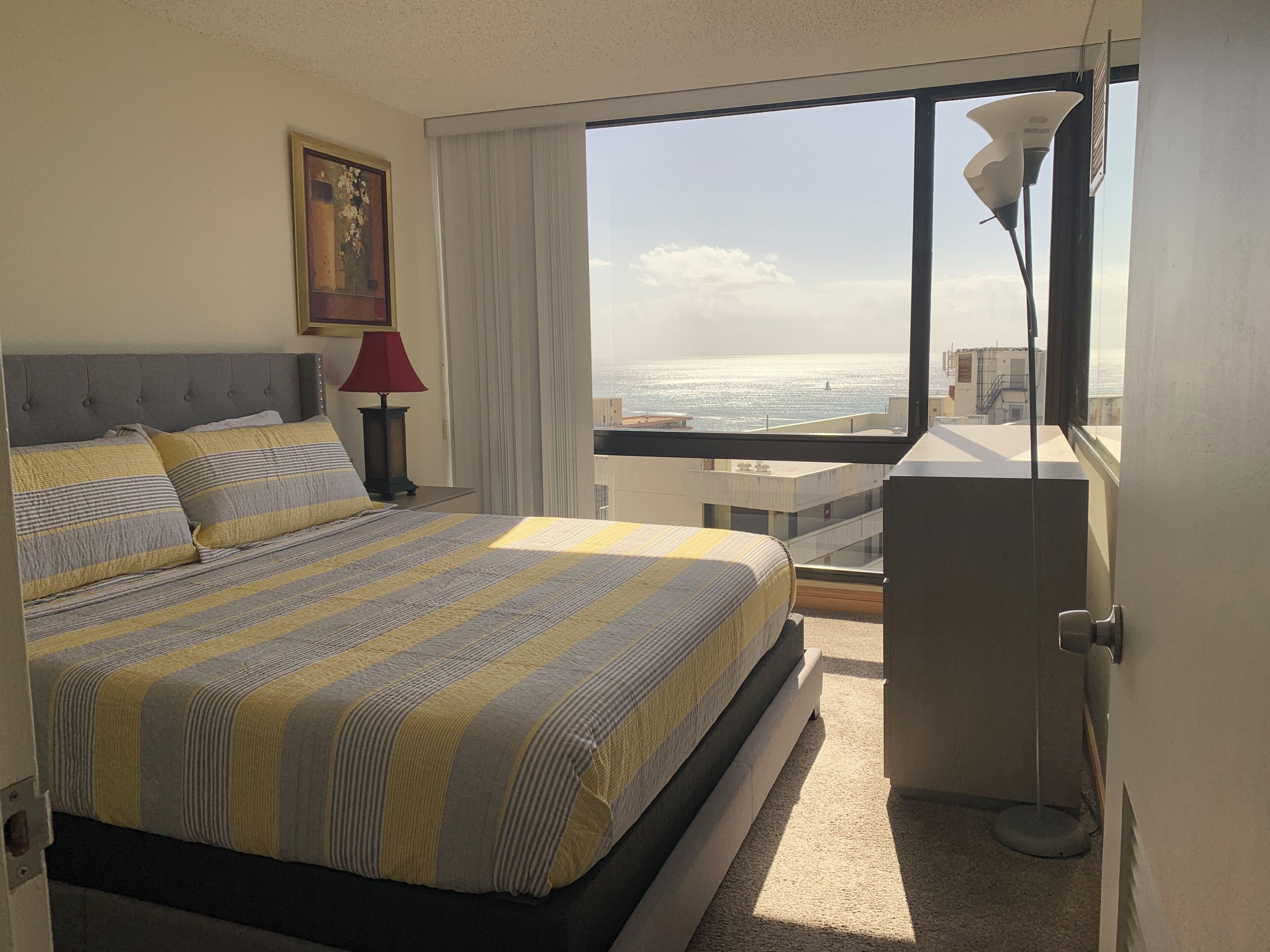 A hotel room with a bed, a nightstand, a lamp, a painting, and a view of the ocean.