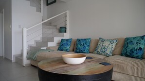 A living room with a beige sofa, blue patterned pillows, a round coffee table with a bowl, and a staircase with a white railing.