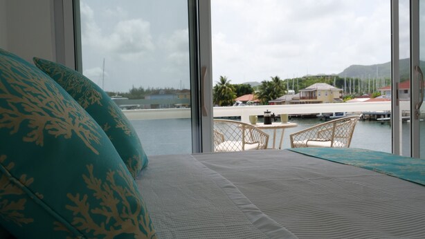 A room with a large window overlooking a marina, a bed with patterned pillows, and a view of the water and distant buildings.