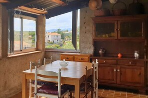 A dining area with a wooden table and chairs, a view of the countryside through large windows, and a wooden cabinet with kitchenware. - Minavita (Pietrosella)
