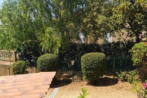 Un jardín ajardinado con diversos árboles y arbustos, una cerca metálica y una zona pavimentada con un canal de drenaje.