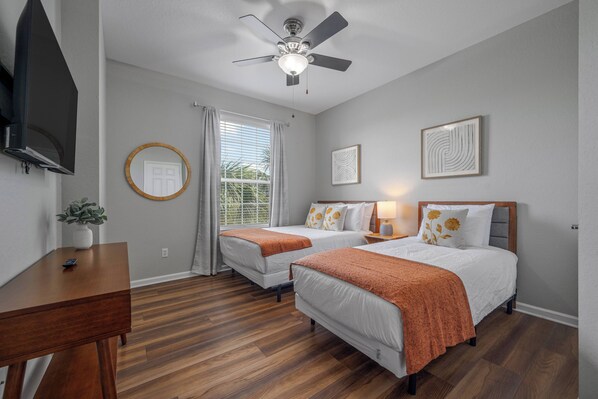 A hotel room with two beds, a ceiling fan, a TV, a desk, and a window with curtains.