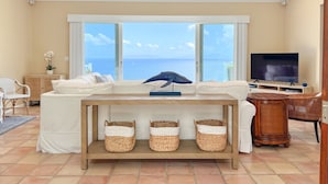 A living room with a white sofa, a wooden coffee table, wicker baskets, a television, and a view of the sea through large windows. - Luxurious Villa w/ Stunning Ocean Views & Pool (St. Thomas)