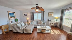 A living room with a striped sofa set, a coffee table, a dining table with chairs, a ceiling fan, and a window with curtains.