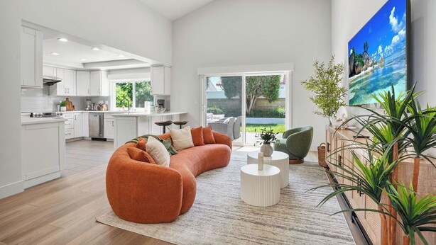 Eine moderne Küche mit weißen Schränken, einem orangefarbenen, gebogenen Sofa und einem großen Fenster mit Blick auf einen Garten.