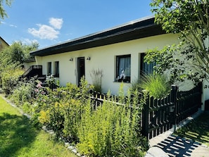Une maison de plain-pied, avec une façade blanche et un toit noir. On peut voir deux fenêtres, l’une avec un rideau et l’autre avec un store. Une clôture noire entoure la propriété, et il y a un jardin avec différentes plantes et fleurs.