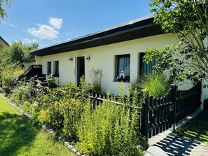 A single-story house with a white exterior and a black roof. There are two visible windows, one with a curtain and one with a blind. A black fence surrounds the property, and there is a garden with various plants and flowers. - F-1112 Vacation apartment Lobbe - Holiday apartment Lobbe F-1112 - house close to the beach (Mönchgut)
