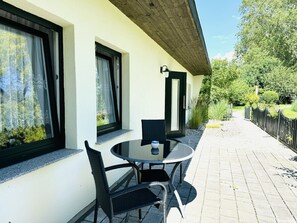 An outdoor seating area with a table and chairs, a window with a curtain, and a pathway leading to a wooden fence. - F-1112 Vacation apartment Lobbe - Holiday apartment Lobbe F-1112 - house close to the beach (Mönchgut)