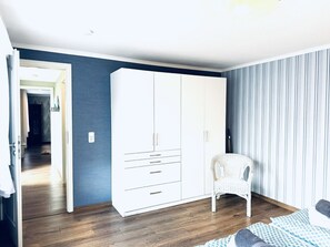 A bedroom with a large white wardrobe, a wicker chair, and a bed with patterned bedding. - F-1112 Vacation apartment Lobbe - Holiday apartment Lobbe F-1112 - house close to the beach (Mönchgut)