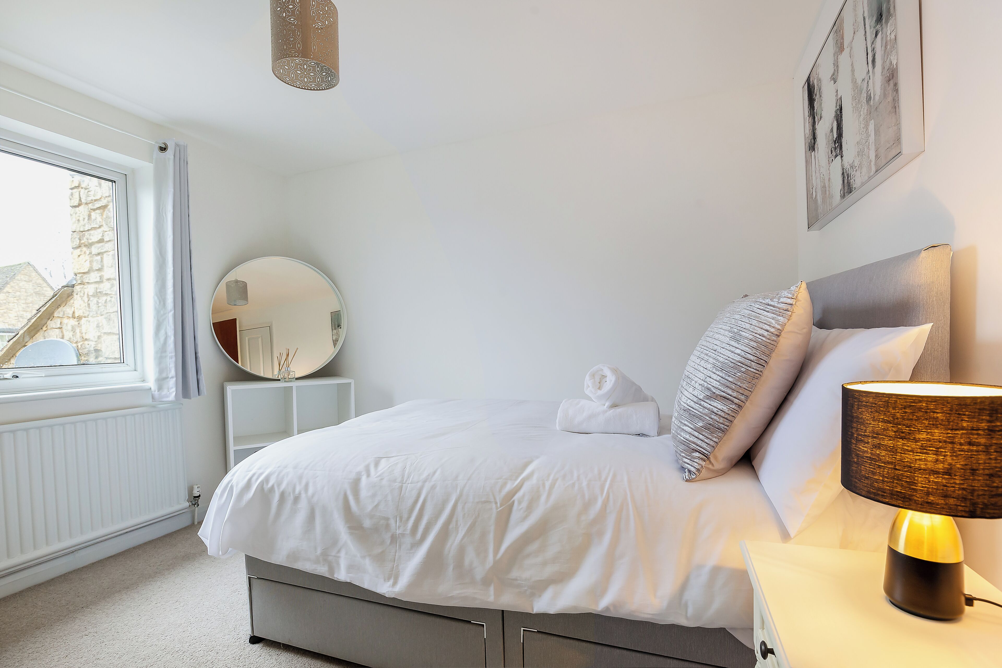 Een net opgemaakt bed met witte lakens, een bedlampje en een ronde spiegel op een witte nachtkastje.