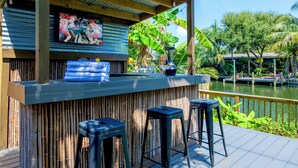 A bar area with a bamboo backdrop, a television screen, and a view of a water body. - 4BR Waterfront Retreat - Private Dock, Kayaks, Hot Tub & Tiki Bar  Walk to Beach (Cocoa Beach)