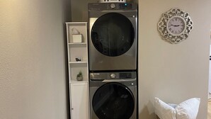 Un cuarto de lavandería con dos lavadoras de carga frontal apiladas, un pequeño armario blanco, un reloj de pared decorativo y una cesta de mimbre con una almohada.