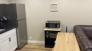 Cocina compacta con refrigerador, microondas y cafetera. Una mesa de madera está colocada en el centro, y un sofá de cuero marrón está apoyado en la pared.