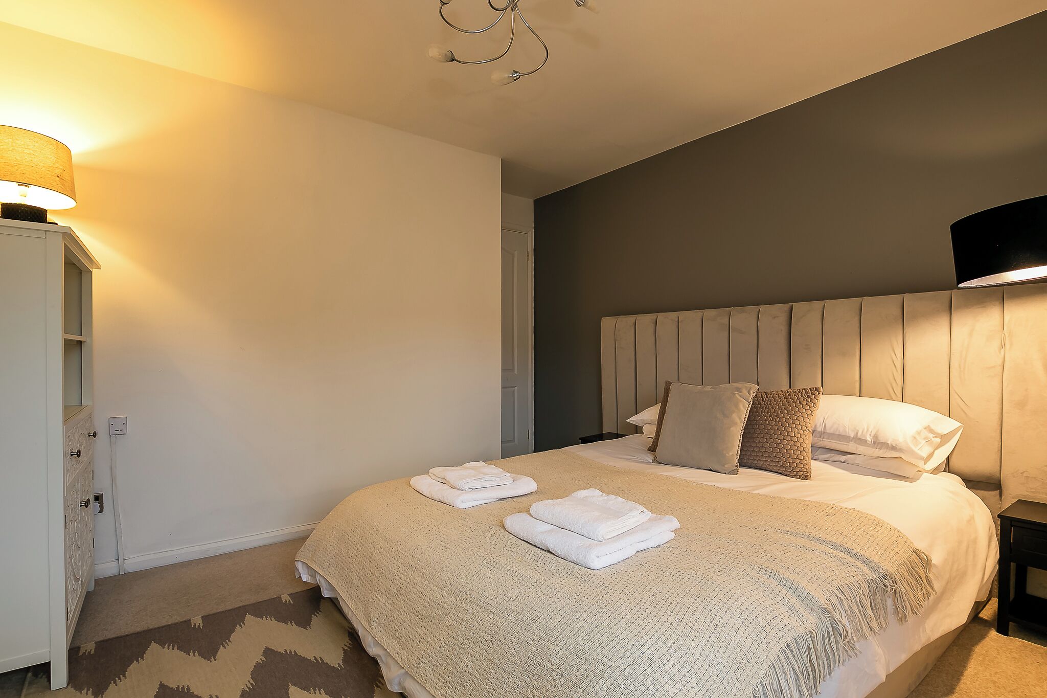 A neatly made bed with folded towels, a bedside table with a lamp, and a wall-mounted light fixture.