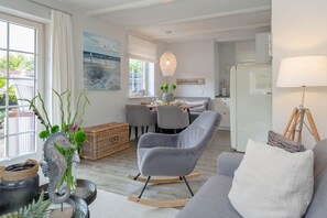 Ett modernt vardagsrum med en grå soffa, en gungstol och en matplats med ett bord och stolar. Det finns ett stort fönster, en vägglampa och en dekorativ skulptur föreställande ett sjöhäst.