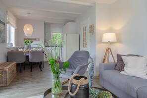 A modern living room with a grey sofa, a dining area with a table and chairs, and a kitchen in the background. - Salty Premium - Salty Premium - Moderne 2 Zimmerwohnung mit Terrasse (Westerland)