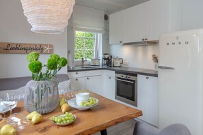 Una cocina moderna con armarios blancos, un horno de acero inoxidable y un refrigerador blanco. Hay una mesa de madera con un florero de plantas verdes y un cuenco de uvas.