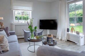 Ett modernt vardagsrum med en grå soffa, ett runt soffbord, en platt-TV och ett stort fönster med utsikt över grönområden.