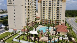 Ein mehrstöckiges Hotel mit Swimmingpool, umgeben von Palmen und einem Parkplatz.