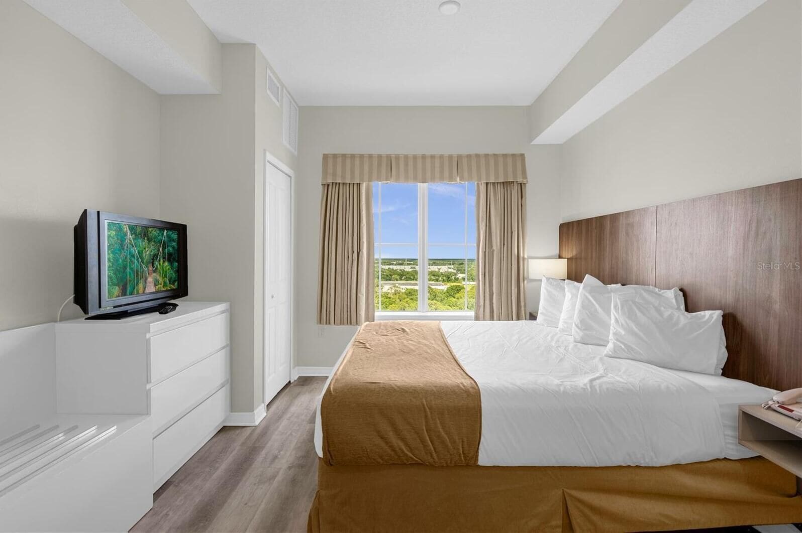 Une chambre d’hôtel avec un lit, un téléviseur posé sur une commode, une fenêtre avec des rideaux et une vue sur la verdure.